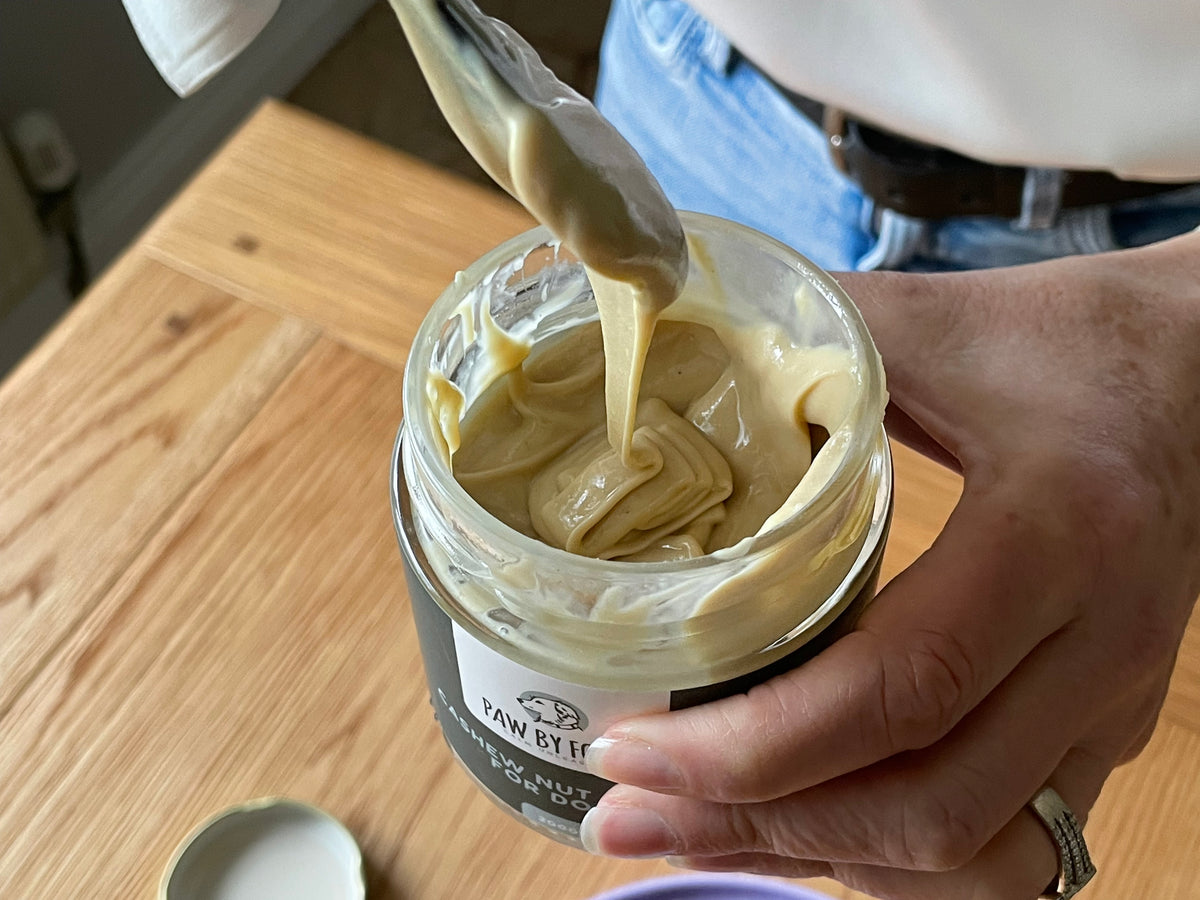 Hand using a spatula to spread cashew nut butter from a Paw by Four jar on a wooden surface