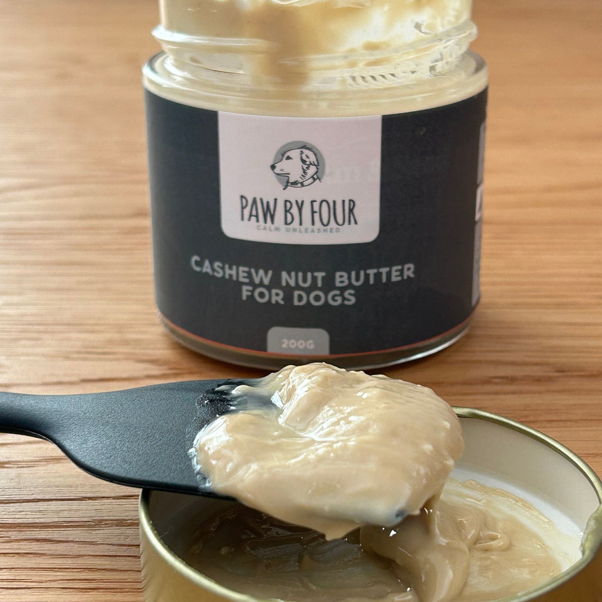 Jar of 'Paw by Four' cashew nut butter for dogs with a spoonful on a wooden surface